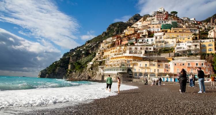 Pebble beach below the colourful cliffside houses of Positano with visitors strolling by the surf.