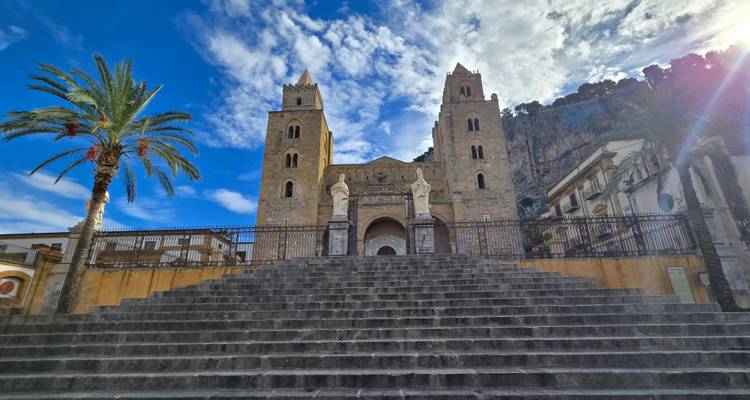 Große steinerne Treppe, die zum Cefalù-Dom führt, flankiert von Statuen und einer hohen Palme unter dramatischem Himmel