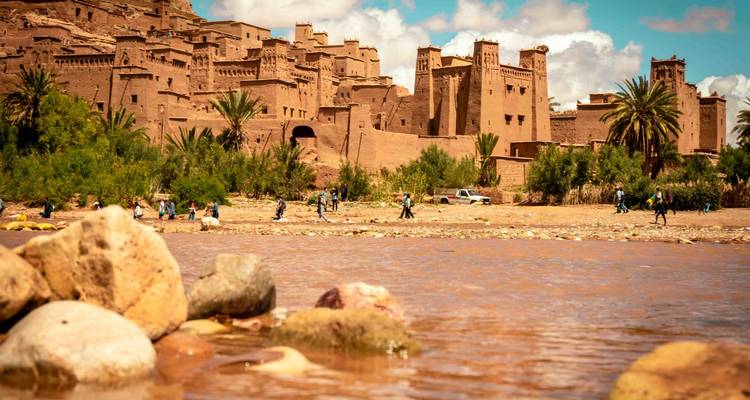 Vue panoramique d'Aït-Ben-Haddou avec rivière et rochers au premier plan.