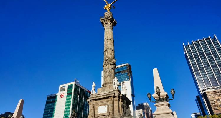 Statue au sommet d'une colonne avec des bâtiments modernes en arrière-plan sous un ciel bleu dégagé.