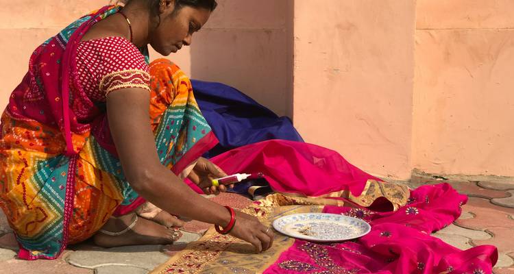A woman in bright traditional clothing embellishes colourful fabric against a peach wall.