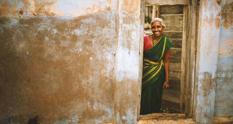 A smiling woman in a green sari stands in the doorway of a weathered village house.