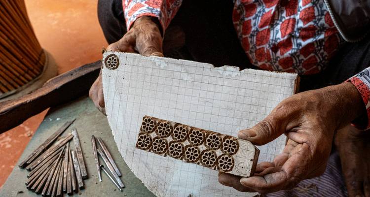 Close-up of an artisan’s weathered hands holding a carved wooden printing block and a tiled design template.