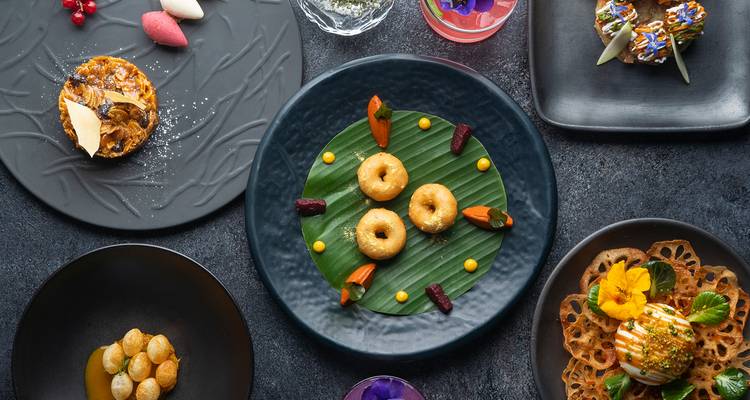 Top-down view of a colourful spread of modern Indian dishes artfully plated on dark ceramic tableware.