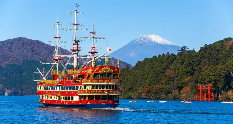Felrode piraten-stijl rondvaartschip vaart over Lake Ashi met de sneeuwbedekte Mount Fuji en beboste heuvels op de achtergrond, en een vermiljoen torii staat aan de oever.