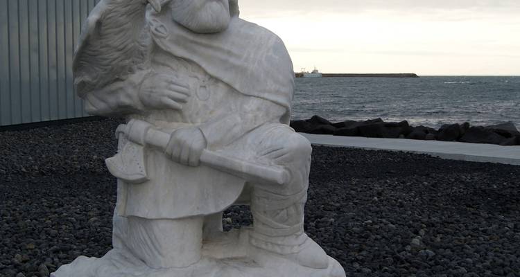 White marble Viking-style statue kneeling with axe beside a rocky seafront under grey skies