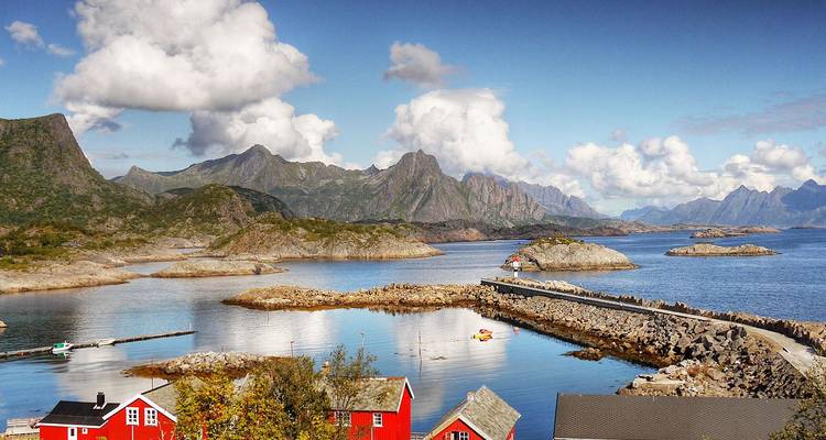 Levendig fjordlandschap met rode kusthuisjes, kalm spiegelend water, rotsachtige eilandjes en dramatische grillige bergen onder verspreide wolken.