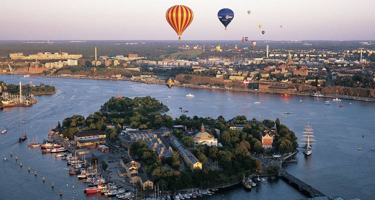 Hot-air balloons drifting above an island-studded Stockholm archipelago with a sprawling cityscape in soft evening light.