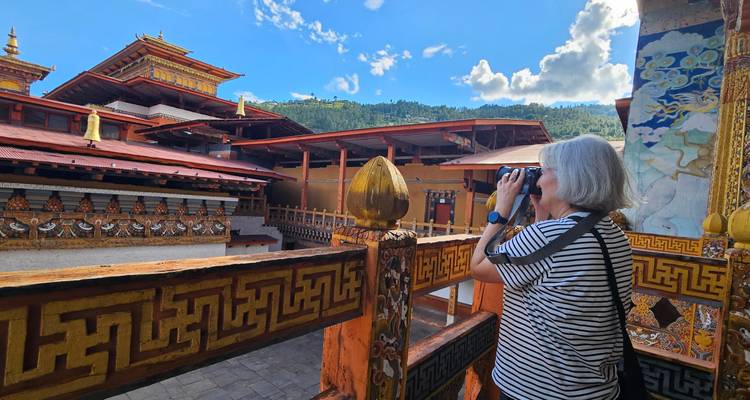 Senior female traveler looks through binoculars at an ornate Bhutanese monastery courtyard under a blue sky.