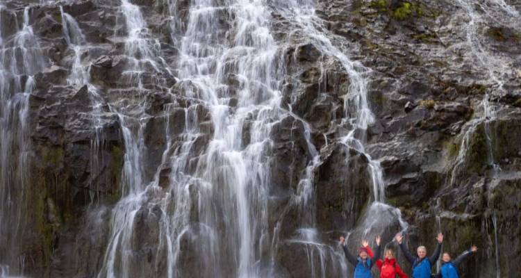 Powerful waterfall cascades down dark basalt cliffs while a group of hikers cheer at its base.