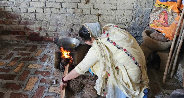 Una mujer se inclina para atender un fuego de leña bajo una estufa de barro dentro de una cocina rústica de ladrillos