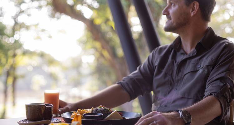A man enjoys a meal and juice at an open-air restaurant overlooking greenery lit by soft morning light.