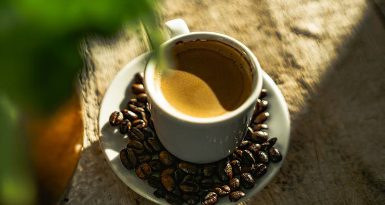 A fresh espresso in a white cup surrounded by roasted coffee beans on rustic wood.