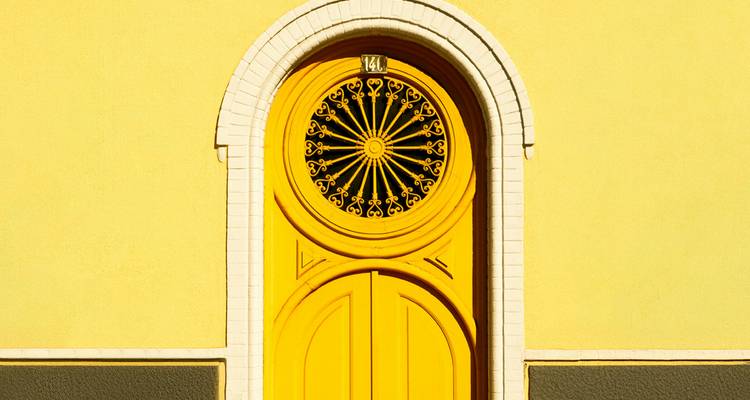 Close-up van een felgeel gewelfd deurtje met een decoratief zwart smeedijzeren raam tegen een gelijkgekleurde muur.