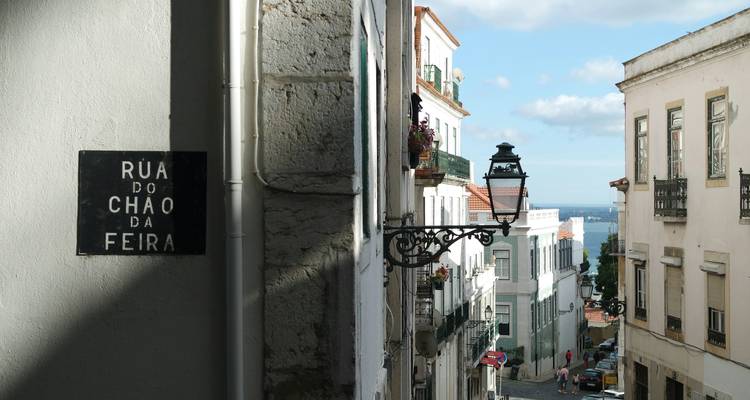 Zonnig straatje in Lissabon met een zwart straatnaambord, een ingewikkeld lantaarnpaal en uitzicht op de blauwe rivier in de verte.
