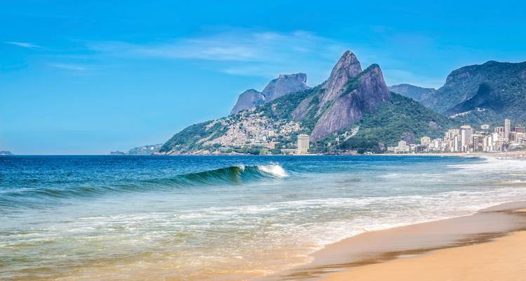 Ruhiger Sandstrand mit sanften Wellen und den Granitgipfeln der Dois Irmãos, die sich hinter der Skyline von Rio erheben.