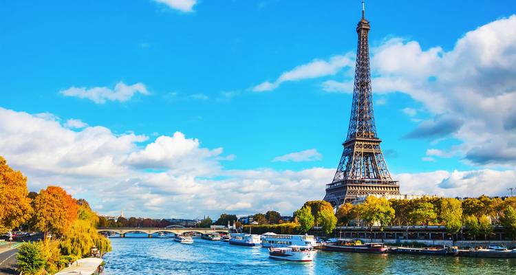 Eiffel Tower rising above the Seine River with boats and autumn trees under a bright blue sky