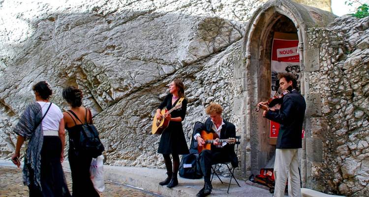 Street musicians performing with guitars and violin beside an old stone wall while pedestrians pass
