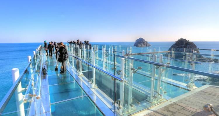 Passerelle de verre avec des touristes, surplombant l'océan et des formations rocheuses en arrière-plan.