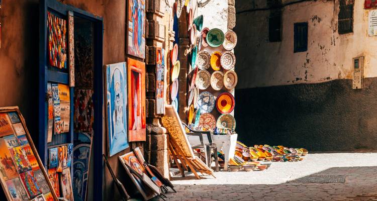 Des peintures colorées et des assiettes en céramique exposées à l'extérieur d'une boutique d'art dans une ruelle marocaine ensoleillée.