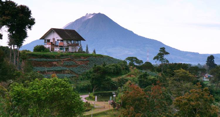 Lonely house perched on a terraced green hillside with a dramatic conical volcano rising in the background.