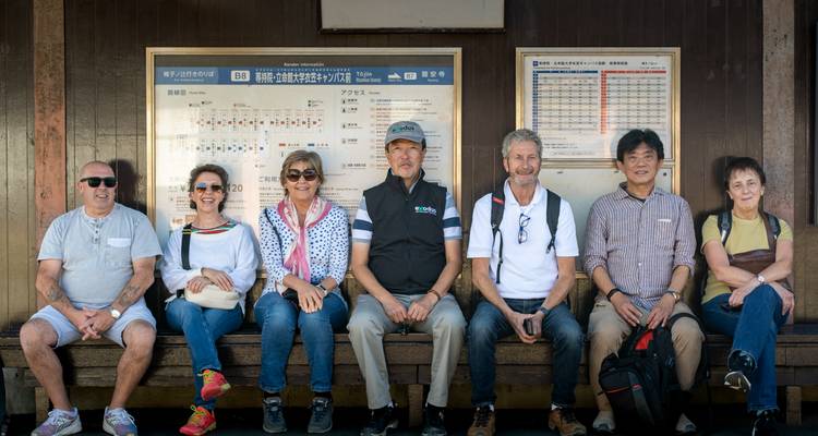 Un groupe de voyageurs est assis sur un banc à une gare rurale sous de grands panneaux d'horaires