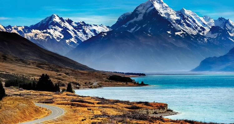 Route sinueuse le long d'un lac turquoise avec des montagnes enneigées.