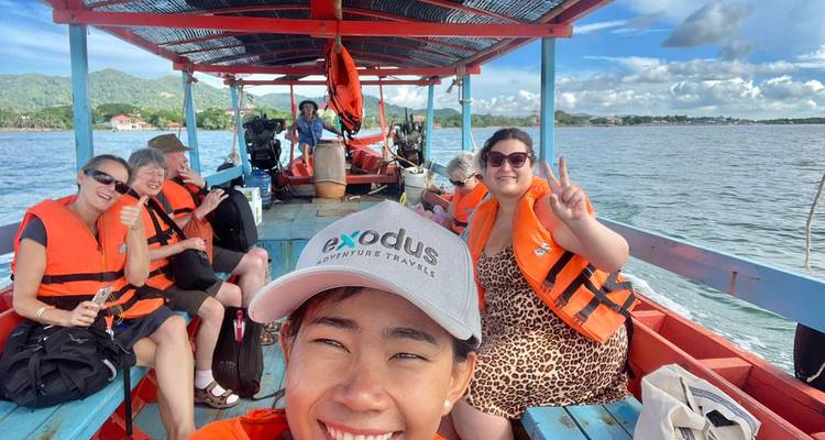 Happy travellers wearing life jackets ride on an open boat across calm water with distant hills.