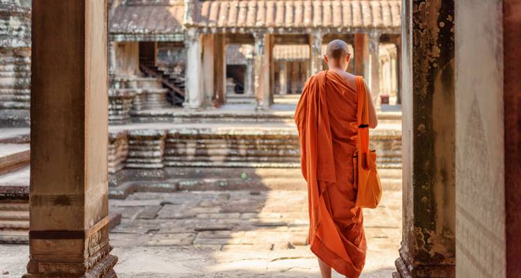 Buddhist monk in orange robes walks through ancient stone columns of a temple complex.