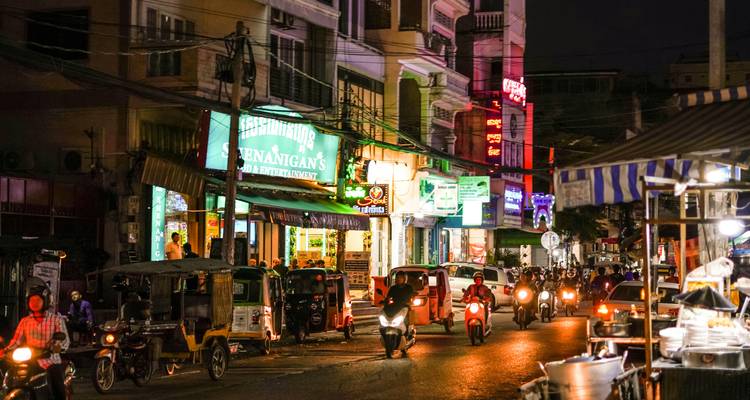 Busy night street in Southeast Asia with neon signs, tuk-tuks and scooters amid lively traffic.