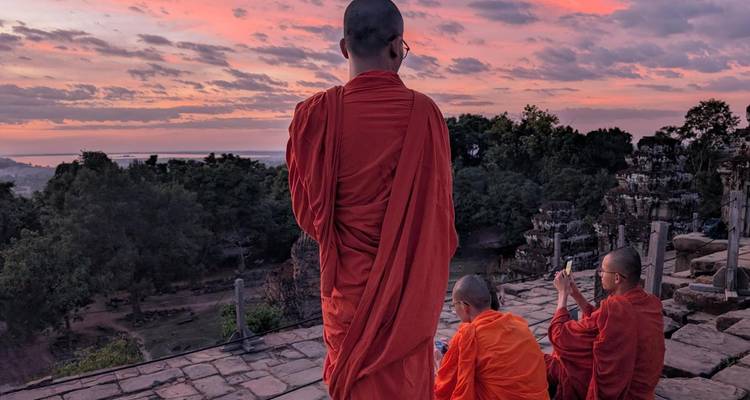 Monks in orange robes watch a pastel sunrise from an ancient stone terrace overlooking forest and distant plains.