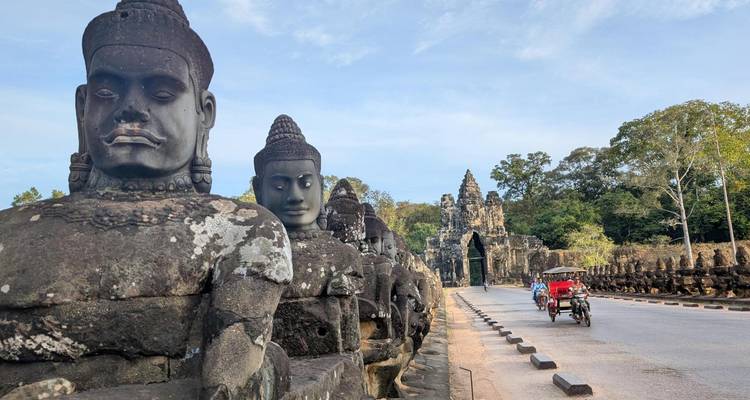Stone guardian statues line the bridge to an ancient Khmer gate while a red tuk-tuk drives past.