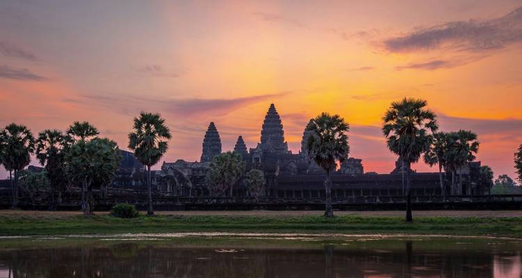 Silhouette of Angkor Wat’s towers framed by palm trees reflected in a still pond under a vivid sunset sky.