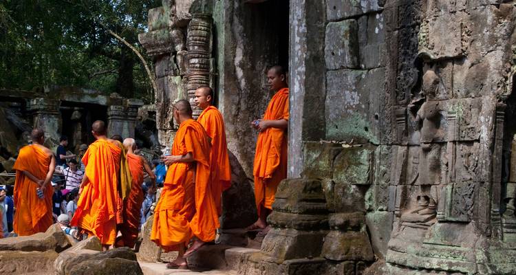 A procession of saffron-robed monks walks through moss-covered temple ruins with intricate bas-reliefs.