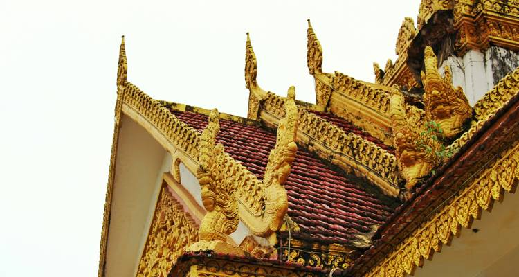Close-up of ornate golden roof carvings and red-tiled gable of a Khmer temple against a white sky.