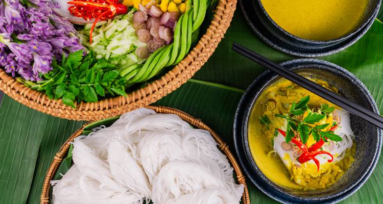 Colorful Cambodian meal with noodles, herbs, curry soup, and fresh vegetables arranged on banana leaves.