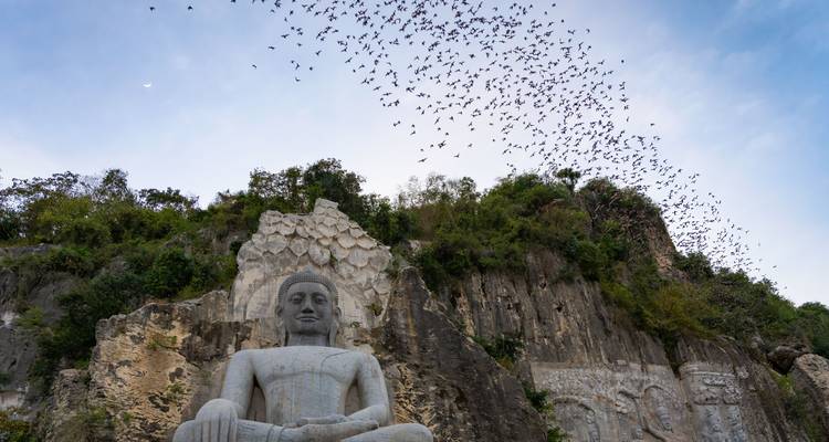 Large seated stone Buddha carved into a cliffside below a swirling flock of bats in the blue evening sky.