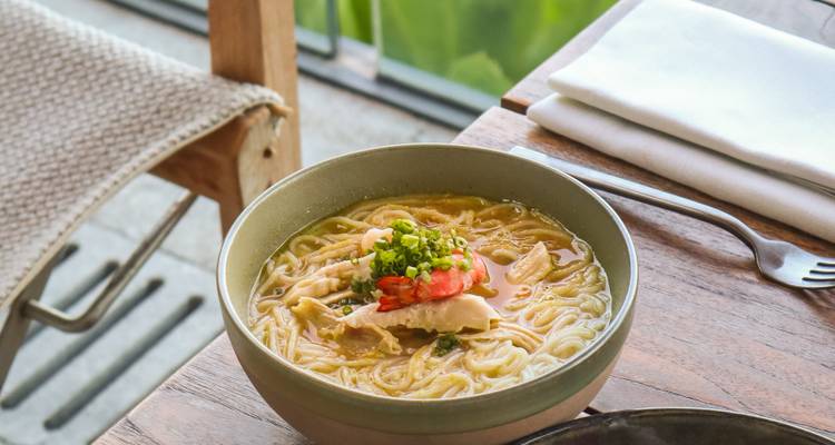 Bowl of steaming noodle soup garnished with herbs and chili on a rustic wooden table beside a window.