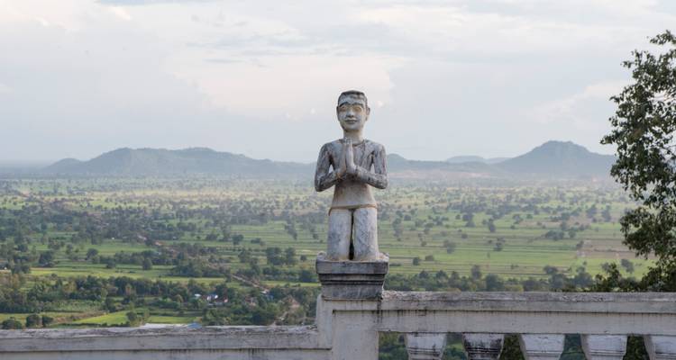 Statue de pierre usée dans une pose de prière surplombant de vastes plaines rurales et des collines lointaines.