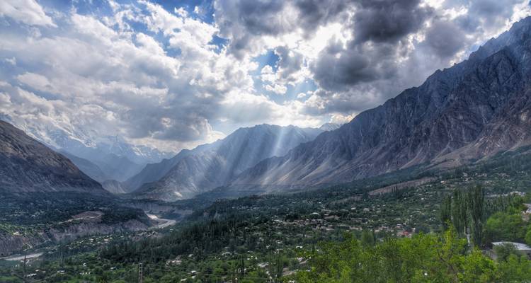 Vue panoramique sur la verte vallée de Hunza avec des rayons de soleil perçant à travers des nuages dramatiques sur les crêtes montagneuses étagées.