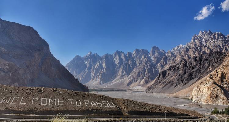 Plaine de vallée désertique avec inscription en pierre 'WELCOME TO PASSU' et les spectaculaires cônes dentelés de Passu qui s'élèvent derrière.