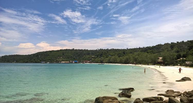 Playa con agua clara y colina boscosa al fondo.