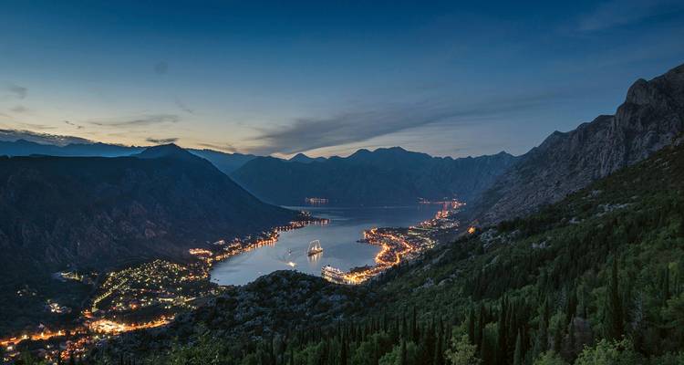 Scenic view of a bay surrounded by mountains at dusk with city lights reflecting on the water.