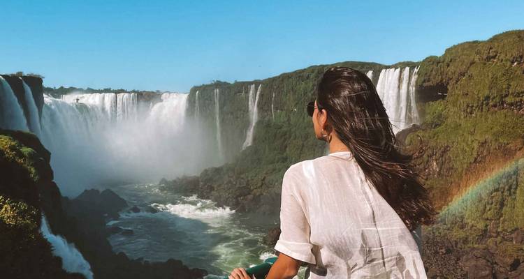 Woman leaning on a railing, looking at waterfalls and a rainbow.