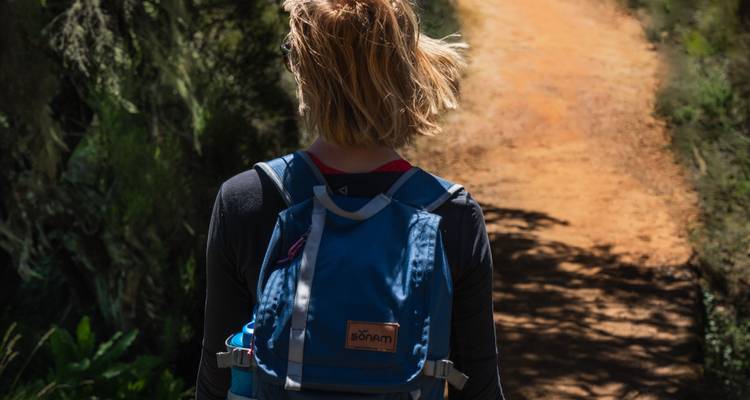 Back view of a hiker with a blue daypack walking along a shaded dirt trail.