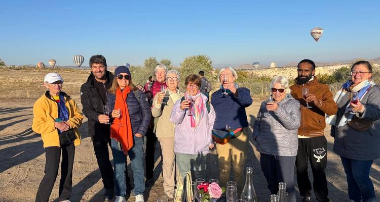 Travel group toasting with drinks in an open field as hot-air balloons rise behind them.
