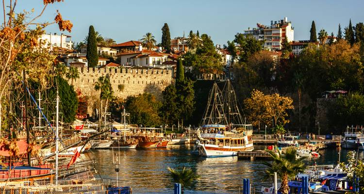 Scenic harbor with historic stone walls, sailboats, and autumn foliage reflected in calm water.