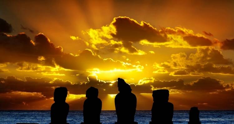 Silhouetted moai figures set against a fiery orange sunset over the Pacific Ocean.
