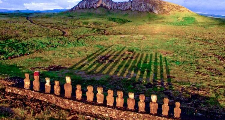 Aerial view of aligned moai casting long shadows across green plains toward a volcanic hill.