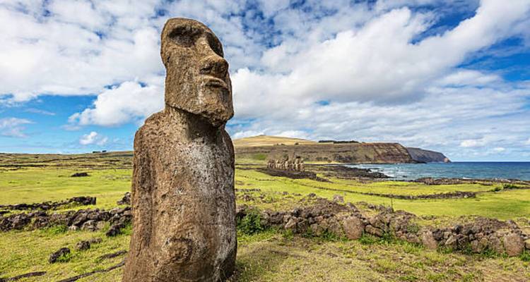 Close-up of a solitary moai statue overlooking grassy cliffs and the sea under partly cloudy skies.